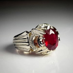 Könnte beinhalten: Ein silberner Ring mit einem großen, ovalen, roten Edelstein, der in einem detaillierten, verzierten Design gefasst ist.