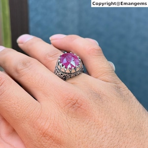 May include: A silver ring featuring a large, oval-cut, deep red gemstone. The ring has an ornate, floral design on the band and a crown-like setting around the stone. The text "Copyright@Emangems" is visible in the top right corner.