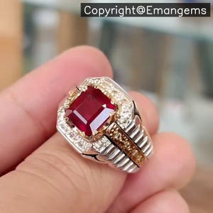 May include: A silver ring featuring a large, square, deep red gemstone. The gemstone is encircled by smaller, clear stones. The ring has a gold band and the text "Copyright@Emangems".