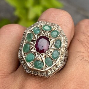 Puede incluir: Un anillo de plata con una gran piedra roja rodeada de piedras verdes más pequeñas. El anillo tiene un diseño detallado con tallas intrincadas.