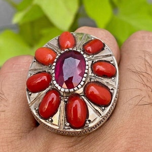 May include: A silver ring with a large, oval-shaped, dark pink gemstone surrounded by smaller, oval-shaped, red coral stones.