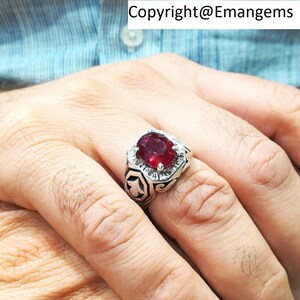 Può includere: Un anello d'argento con una grande gemma rettangolare rosso scuro al centro, circondata da pietre più piccole e trasparenti. L'anello presenta un design geometrico dettagliato sulla fascia. Il testo "Copyright@Emangems" è visibile.