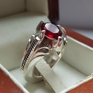 Puede incluir: Un anillo de plata con una gema ovalada de color rojo intenso. El anillo tiene un diseño ornamentado con tallados detallados en la banda y alrededor del engaste de la gema. El anillo se muestra en una caja de joyería.