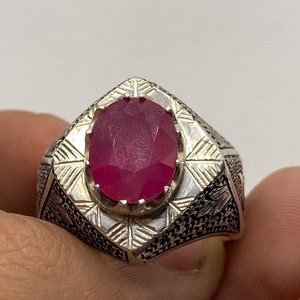 Peut inclure: Une bague en argent avec une pierre précieuse rouge de forme ovale sertie dans une bélière carrée. La bague présente des détails gravés complexes sur la bande.