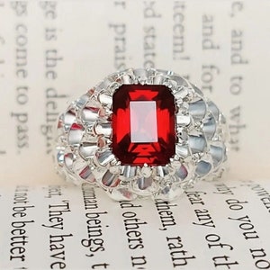Peut inclure: Une bague en argent avec une grande pierre précieuse rectangulaire rouge sertie au centre. La bague a une bande texturée.