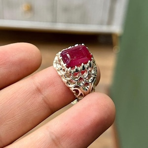 May include: A silver ring with an intricate design features a large, deep red gemstone in the center.