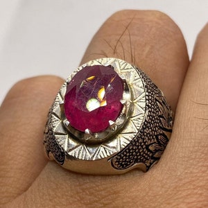 Peut inclure: Une bague en argent avec une grande pierre précieuse rouge de forme ovale sertie dans un décor orné et détaillé.