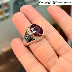 May include: A silver and gold ring featuring a large, oval-cut, deep red gemstone. The ring has a gold setting around the gemstone, accented with small, clear stones. The ring is displayed on a hand.