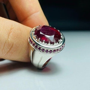 Puede incluir: Anillo de plata con una gran gema de rubí de corte ovalado. El rubí es de color rojo intenso y está rodeado de rubíes redondos más pequeños. El anillo tiene un diseño detallado y ornamentado.