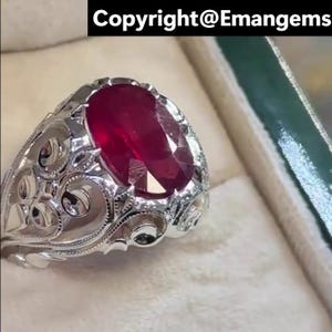 Puede incluir: Anillo de plata con una gema ovalada de color rojo intenso. El anillo presenta un diseño de filigrana ornamentado alrededor de la gema. El texto "Copyright@Emangems" se encuentra en la parte superior de la imagen.