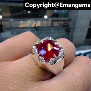 May include: A silver ring with a large, square-cut, deep red gemstone. The gemstone is encircled by smaller, clear stones. The ring is held by a hand, with the text "Copyright@Emangems" visible at the top.