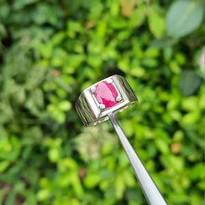 Könnte beinhalten: Ein silberner Ring mit einem großen, oval geformten, roten Edelstein. Der Ring hat eine rechteckige Fassung, in der der Edelstein in einer Zargenfassung gefasst ist. Der Hintergrund ist verschwommen grün.