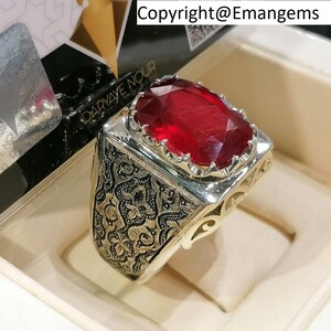 Puede incluir: Un anillo de plata con un diseño intrincado y una gran piedra roja de forma ovalada. El anillo está engastado en una banda de plata con un borde decorativo alrededor de la piedra preciosa.