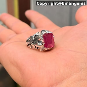 May include: A silver ring with an elaborate design, set with a large, rectangular, deep red gemstone. The ring is displayed in a hand, highlighting the detailed metalwork and the vivid colour of the stone. The text "Copyright@Emangems" is visible.