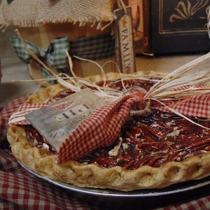 May include: A freshly baked pecan pie on a metal pie plate, with a flaky crust and a filling of pecans in a sweet glaze. The pie is decorated with a red and white checkered fabric and a tag that says "Pecan Pie".