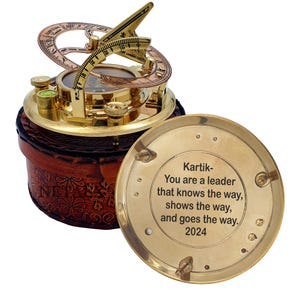 Peut inclure: Un cadran solaire en laiton avec une boussole, présenté dans un étui en cuir. Le cadran solaire comporte un gnomon et un cadran circulaire avec des chiffres romains. Le couvercle de l'étui porte l'inscription "Kartik - You are a leader that knows the way, shows the way, and goes the way. 2024."