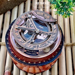 Puede incluir: Una brújula de latón con una funda de cuero. La brújula tiene un reloj de sol y una aguja de brújula. La funda es marrón con un patrón decorativo. La brújula está etiquetada "Gilbert & Sons London (1895)".