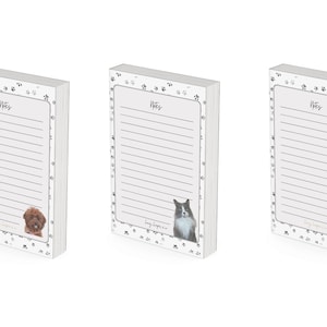 May include: Three white notepad sets with black paw print designs. Each notepad has a different animal on the bottom: a brown dog, a black and white cat, and a white dog.