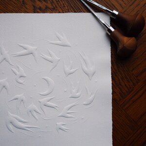 May include: A white paper print featuring a flock of embossed swallows, a crescent moon, and stars. Two wood-handled carving tools rest on a dark wood surface, suggesting a printmaking process. The artwork has a textured, tactile quality.