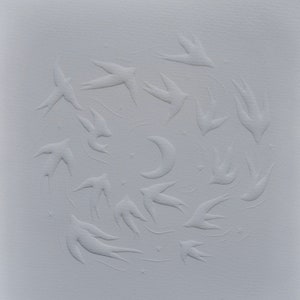 May include: A white paper print featuring a flock of birds in flight, arranged in a circular pattern around a crescent moon and stars. The birds and celestial bodies are embossed, creating a textured effect.