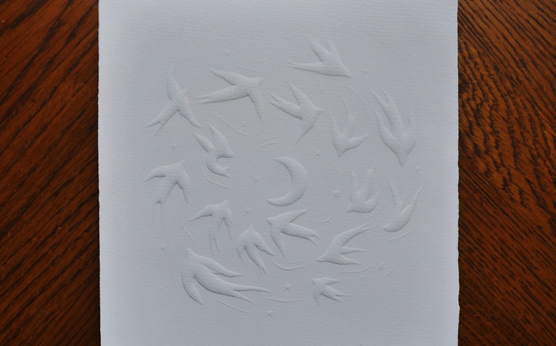 May include: A white paper print featuring a flock of birds in flight, arranged in a circular pattern around a crescent moon and stars. The birds and celestial bodies are embossed, creating a textured effect.