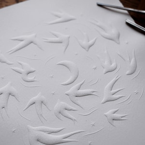 May include: A white paper with an embossed design featuring a flock of swallows, a crescent moon, and stars. The artwork is created with a relief technique, giving the image a three-dimensional effect. The paper has a textured surface.
