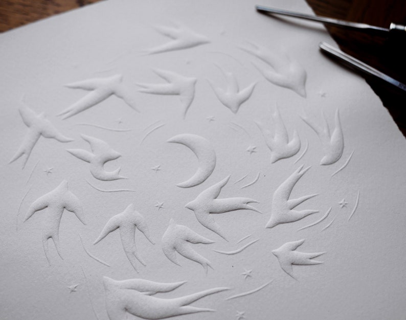 May include: A white paper with an embossed design featuring a flock of swallows, a crescent moon, and stars. The artwork is created with a relief technique, giving the image a three-dimensional effect. The paper has a textured surface.