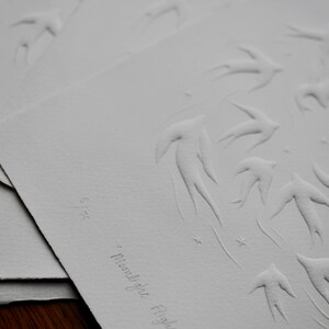 May include: A series of white paper prints featuring embossed designs of birds in flight, a crescent moon, and stars. The phrase "Moonlight Flight" is written in cursive. The print is numbered 5/75.