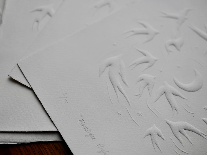 May include: A series of white paper prints featuring embossed designs of birds in flight, a crescent moon, and stars. The phrase "Moonlight Flight" is written in cursive. The print is numbered 5/75.
