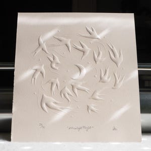 May include: A cream-colored print featuring a circular design of embossed birds in flight around a crescent moon and stars. The text "Moonlight Flight" is below the image, along with the edition number 26/75.