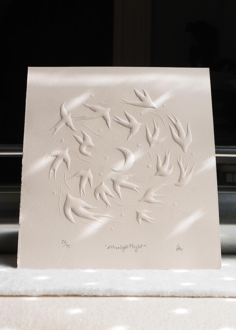 May include: A cream-colored print featuring a circular design of embossed birds in flight around a crescent moon and stars. The text "Moonlight Flight" is below the image, along with the edition number 26/75.