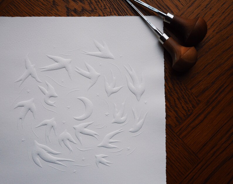 May include: A white paper print featuring a flock of embossed swallows, a crescent moon, and stars. Two wood-handled carving tools rest on a dark wood surface, suggesting a printmaking process. The artwork has a textured, tactile quality.