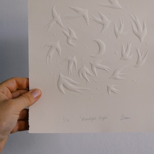 May include: A cream-colored print featuring embossed swallows in flight, a crescent moon, and stars. The artwork is titled "Moonlight Flight" and is numbered 5/75. The artist's signature is visible.