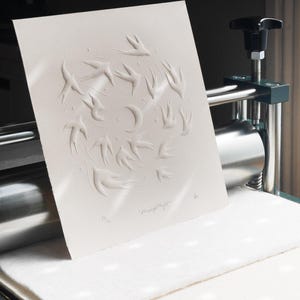 May include: A white paper print with an embossed design of birds in flight around a crescent moon. The artwork is titled "Moonlight Flight" and is numbered 35/75. The print is on a press.