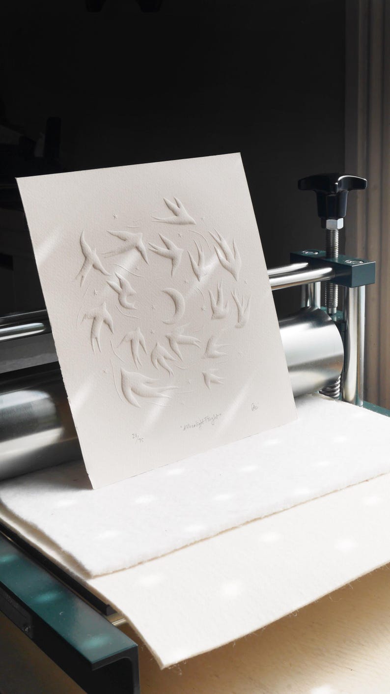 May include: A white paper print with an embossed design of birds in flight around a crescent moon. The artwork is titled "Moonlight Flight" and is numbered 35/75. The print is on a press.