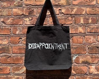 Puede incluir: Bolsa de tela negra con texto blanco que dice "DISAPPOINTMENT" colgada en una pared de ladrillo.