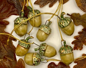 May include: A selection of ceramic acorn ornaments with gold string hangers. The acorns are mainly tan with green and gold detailing. Brown oak leaves surround the ornaments, creating an autumnal theme.