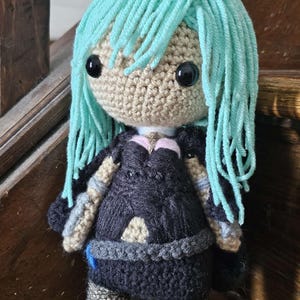 Puede incluir: Muñeca de ganchillo hecha a mano con cara marrón claro, ojos negros y cabello de hilo turquesa. La muñeca viste un vestido negro con detalles grises y beige. La muñeca está sobre una superficie de madera.