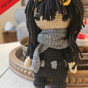 May include: Crocheted doll with black yarn hair, a gray scarf, and a dark gray outfit. The doll has large black eyes and small yellow accents in its hair. The text "PDF PATTERN ONLY" is in the upper left corner.