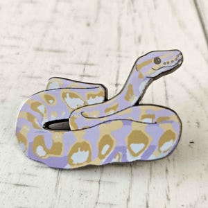 May include: A colorful snake-shaped pin with a light purple base and tan and white spots. The pin is a cartoon-style illustration of a snake, with a curved body and a raised head. The pin is set against a white wooden background.