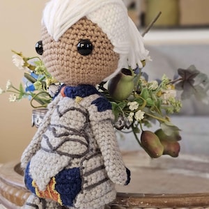 Może przedstawiać: Figurka amigurumi wykonana na szydełku przedstawiająca osobę z białymi włosami, ubraną w szary pancerz z niebieskimi i pomarańczowymi akcentami. Figurka stoi na drewnianej tacy z kwiatową aranżacją z tyłu.