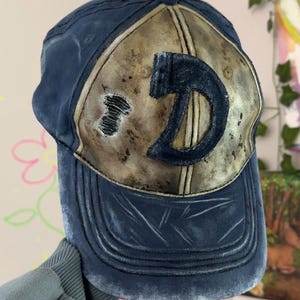 May include: A navy blue baseball cap with a distressed finish and a faded tan front panel. The cap features a large, dark blue letter D patch centered on the front, along with a small, frayed hole detail on the left side of the tan panel.