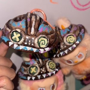 Puede incluir: Llaveros de criaturas de cerámica pintados a mano, marrones, con detalles en azul, rosa y blanco. Cada llavero presenta una cara única con ojos circulares y en forma de cruz. Los llaveros están sujetos a un anillo metálico naranja.