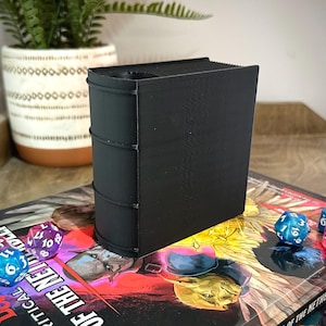 May include: Black book-shaped dice tower with a slot on top for rolling dice. The tower is sitting on a tabletop game book with a fantasy illustration and dice.