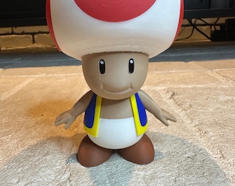 May include: A white and red mushroom-shaped figurine with a brown face and blue and yellow vest. The figurine is standing on a light brown surface.