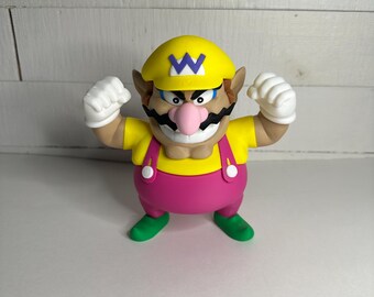 May include: A figurine of the video game character Wario. The figure is wearing a yellow hat with a purple "W" on it, a yellow shirt, pink overalls, and green shoes. His fists are clenched, and he has a large mustache and a pink nose.