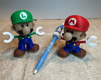May include: Two small plastic figurines of Mario and Luigi characters from the video game Super Mario Bros. The figures are wearing their signature red and green outfits and hats. The figures are holding a blue pen with the word "ABC" printed on it.