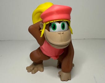 May include: A figurine of a cartoon monkey in a crouching pose. The monkey has brown fur, a tan face, and green eyes. It wears a pink shirt and a red hat with yellow hair. The figurine is on a white surface.