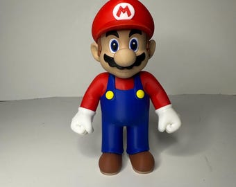May include: A figurine of the video game character Mario. The figure is wearing a red hat with a white "M", a red shirt, blue overalls with yellow buttons, white gloves, and brown shoes. The figure has a black mustache and blue eyes.