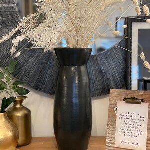 Könnte beinhalten: Eine schwarze Keramikvase mit einer breiten Öffnung und einer schmalen Basis, gefüllt mit getrocknetem weißen Pampasgras und Farnen. Die Vase steht auf einem Holztisch neben einer goldenen Metallvase und einem gerahmten Zitat, das lautet: "Seien wir dankbar für die Menschen, die uns glücklich machen: Sie sind die charmanten Gärtner, die unsere Seelen zum Blühen bringen. Marcel Proust"